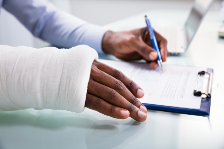 Image of a person's hands completing a form on a clipboard. None of the text on the form can be read. The person has a cast on their right hand.